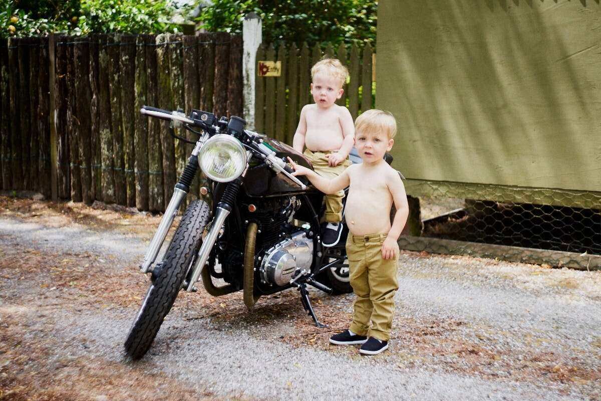 Two toddler boys posing shirtless beside a vintage black motorcycle on a gravel path outdoors.