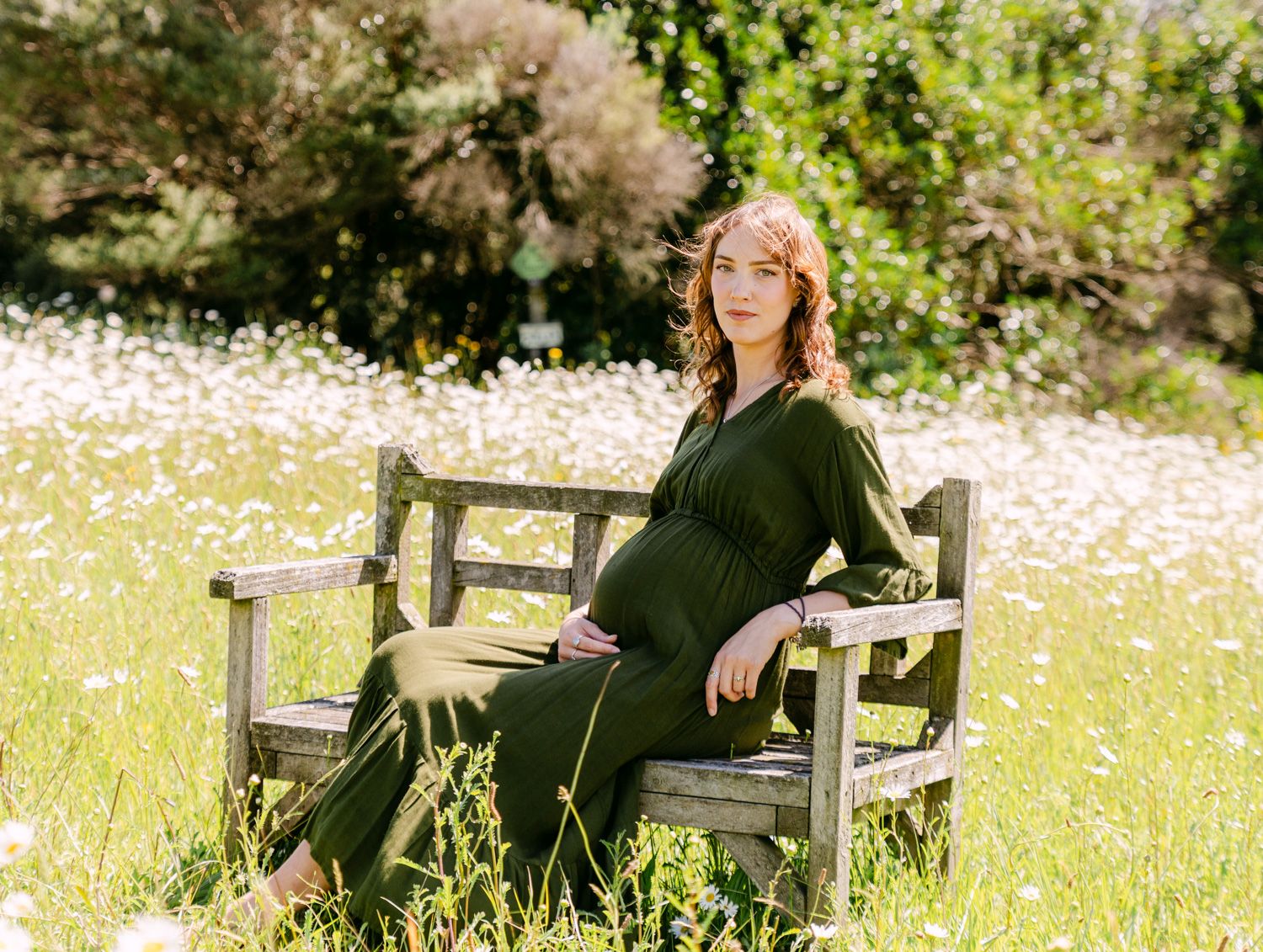 Pregnant woman in olive green dress sitting on rustic wooden bench in wildflower meadow with lush greenery backdrop.