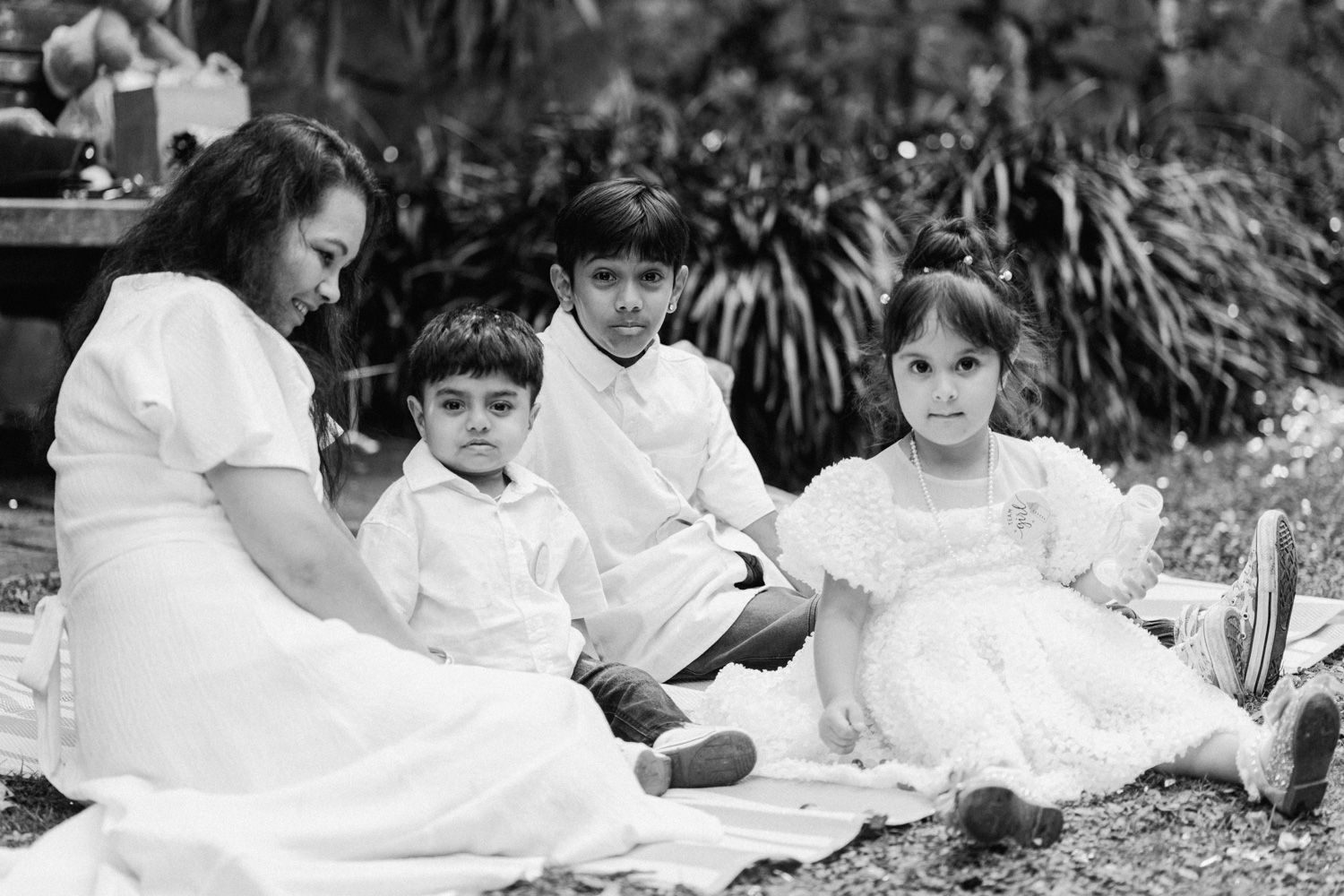 Black and white photo of a woman and three children in white outfits sitting on a blanket outdoors.