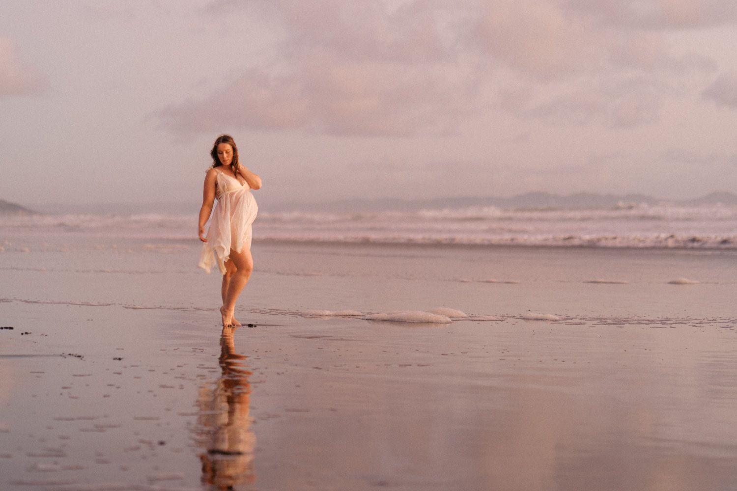 A woman in a white dress walks barefoot along a serene beach at sunset with pink skies.