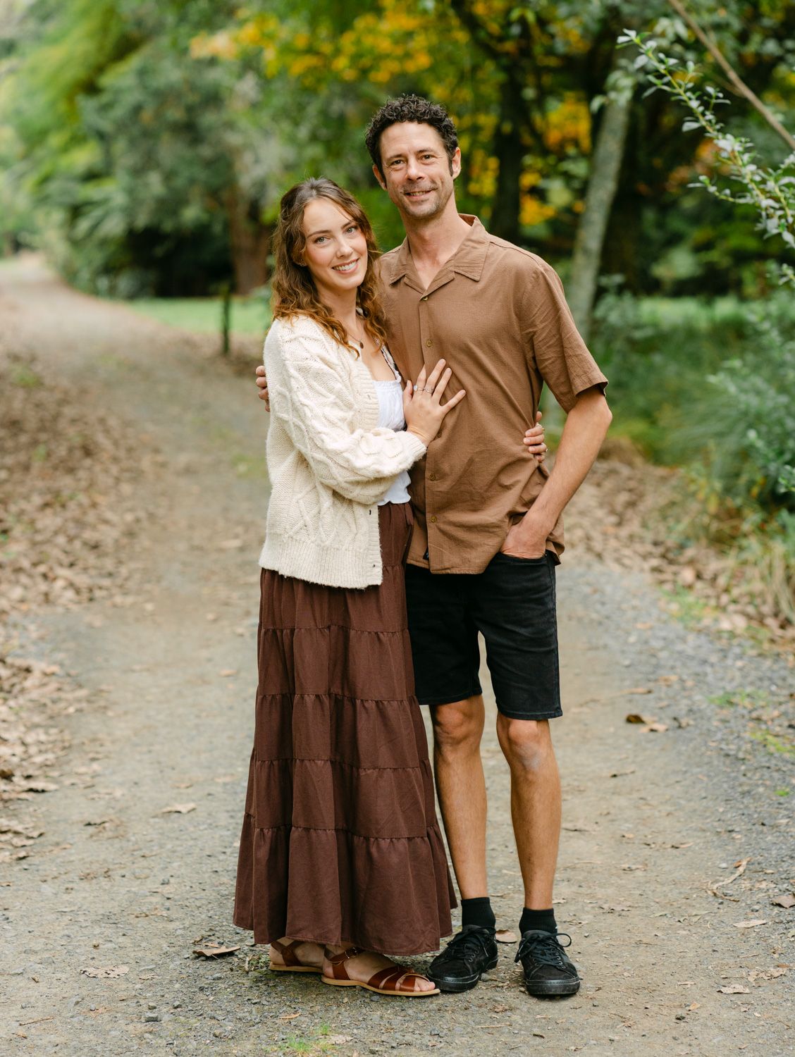 A couple smiles together on a scenic forest path, woman in cream top and brown skirt, man in tan shirt and black shorts.