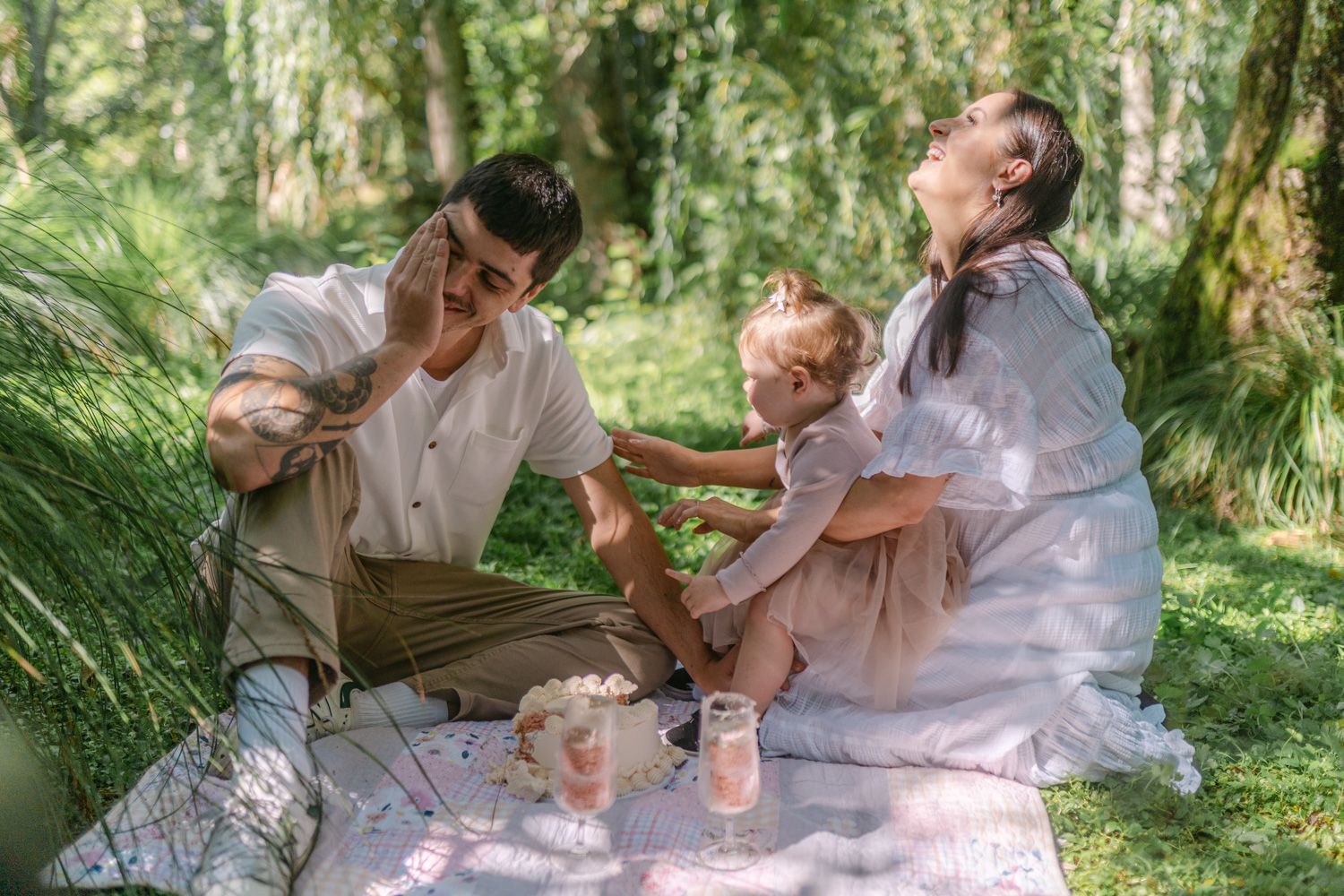 Family enjoying a sunny outdoor picnic on a blanket in a lush green forest setting.