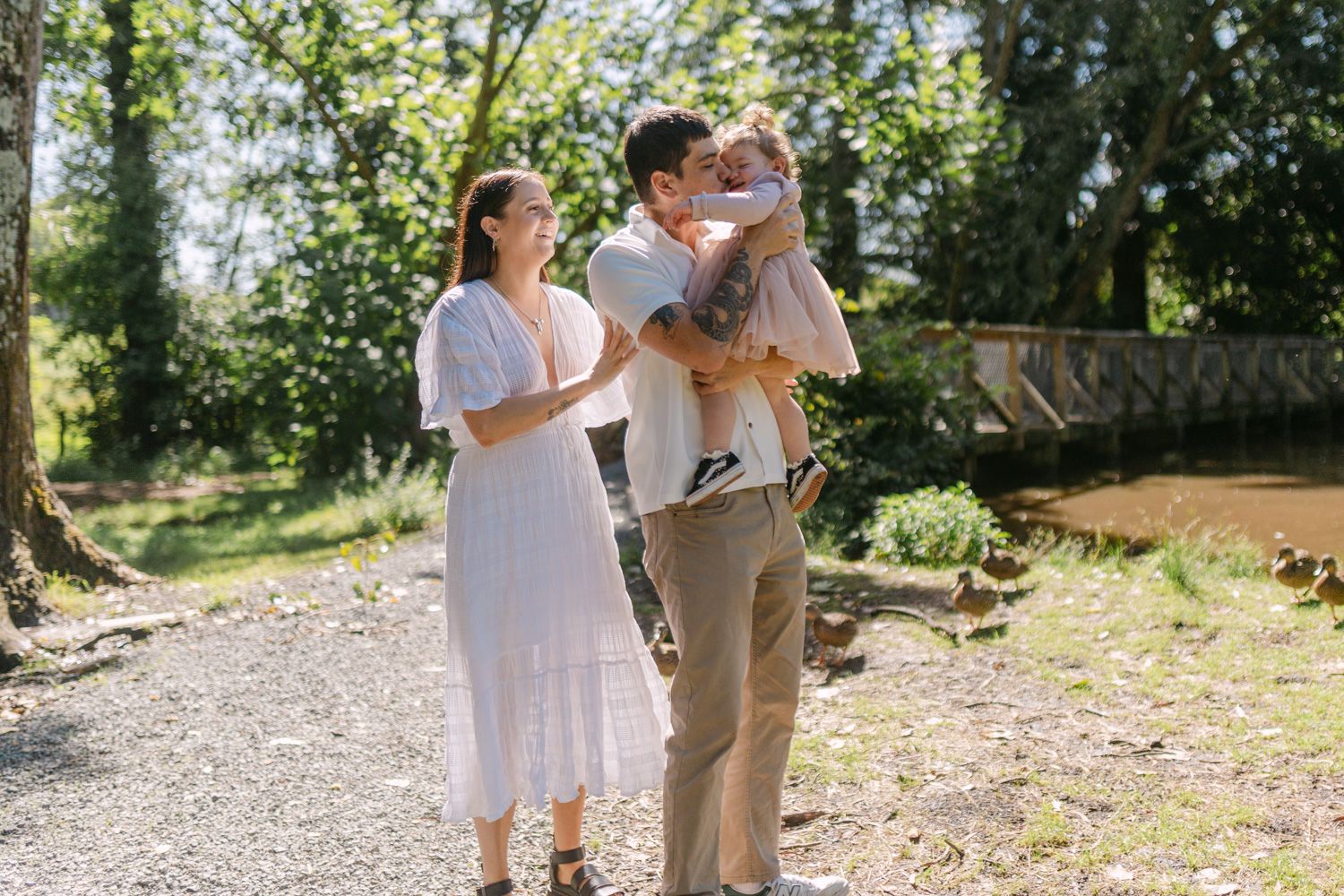 A family of three enjoys a sunny outdoor walk, with a man kissing a baby while a woman in a white dress smiles nearby.