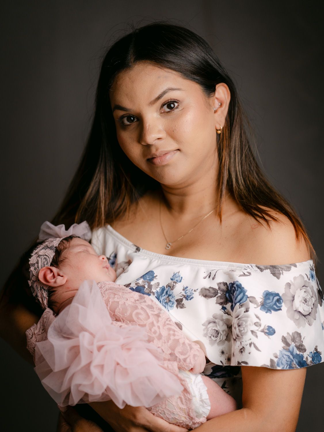 A young woman in a floral off-shoulder top holds a sleeping newborn baby dressed in a pink lace outfit with a bow headband.