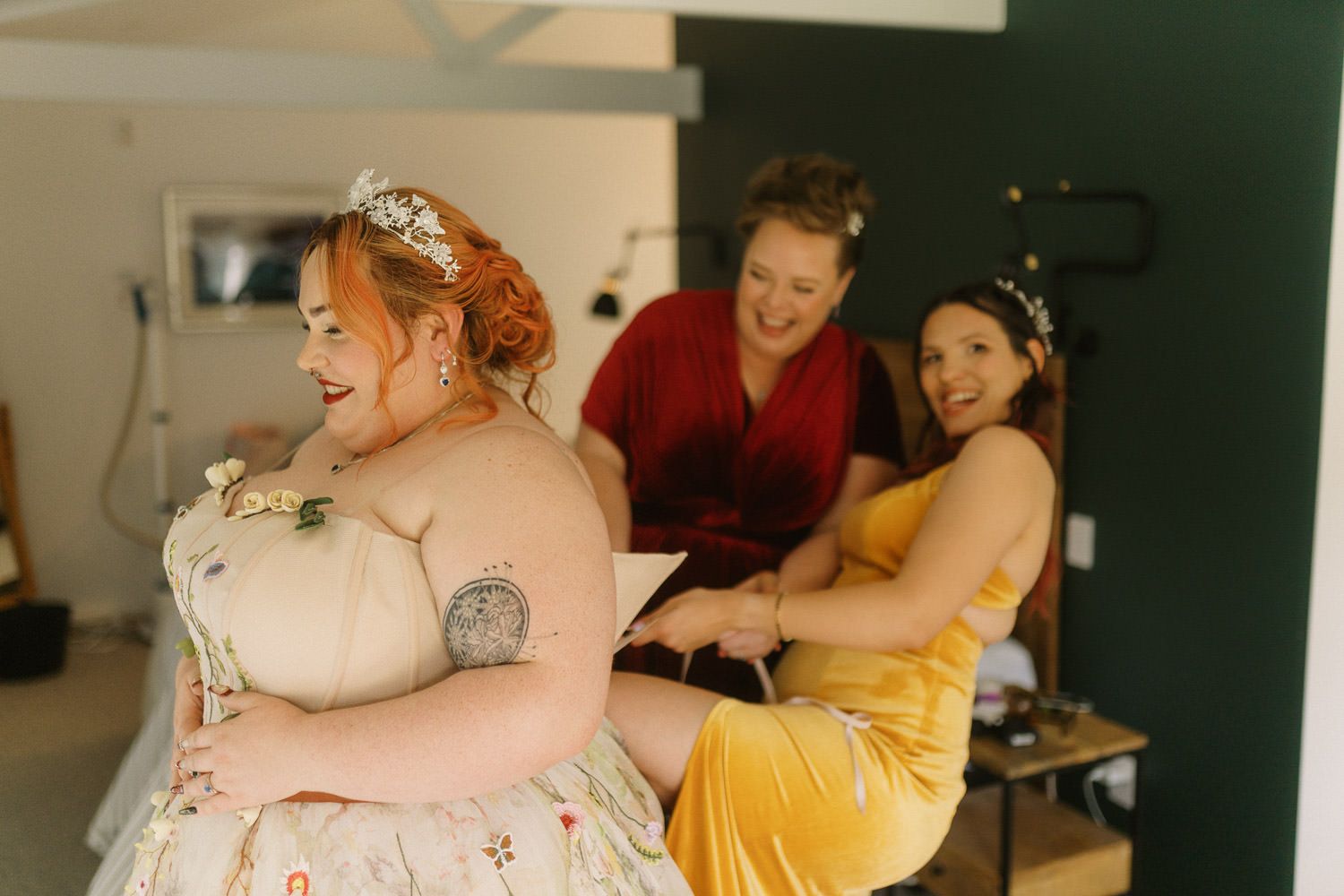 Bride laughing joyfully as bridesmaids help her into her floral wedding dress before the ceremony.