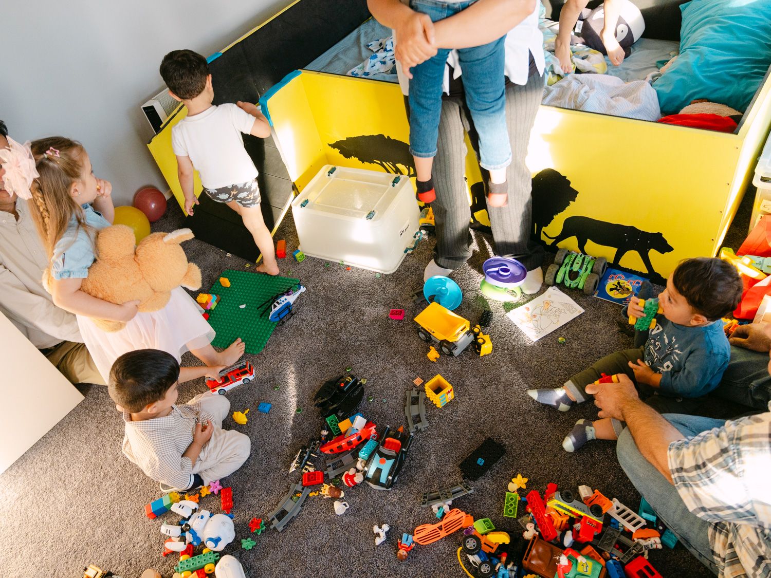 Children playing with colorful Lego toys scattered on floor in a playroom, viewed from above.