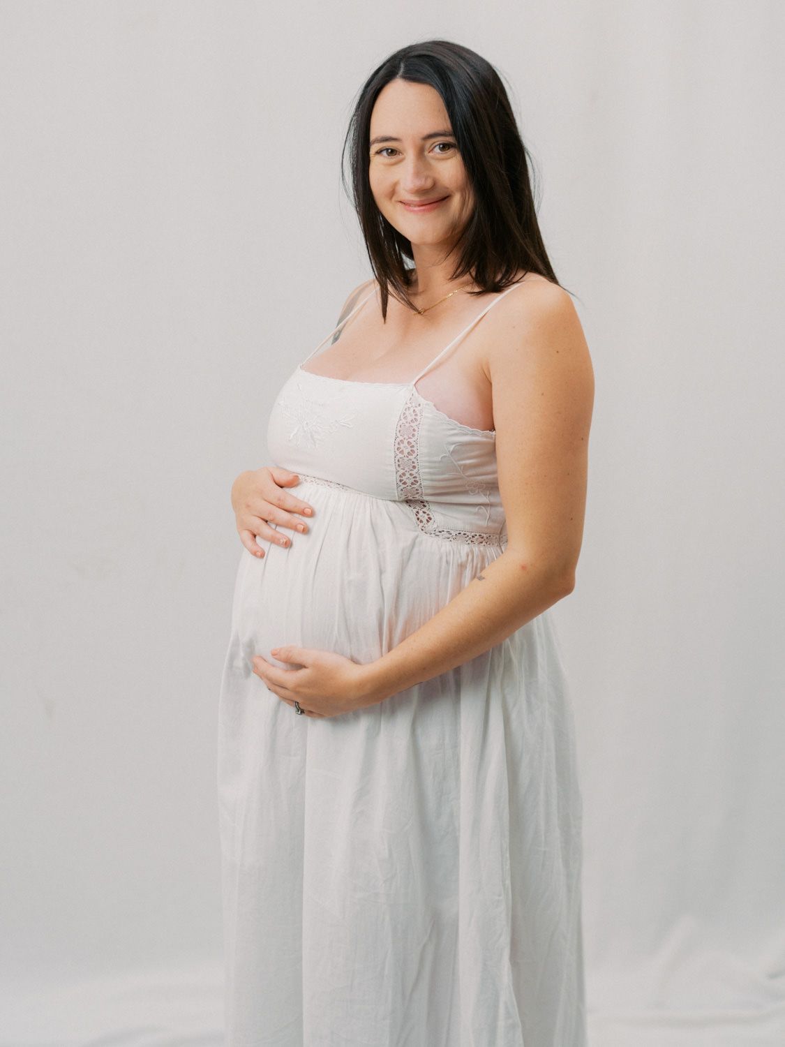 Pregnant woman in white dress cradling baby bump during maternity photo session against light background.