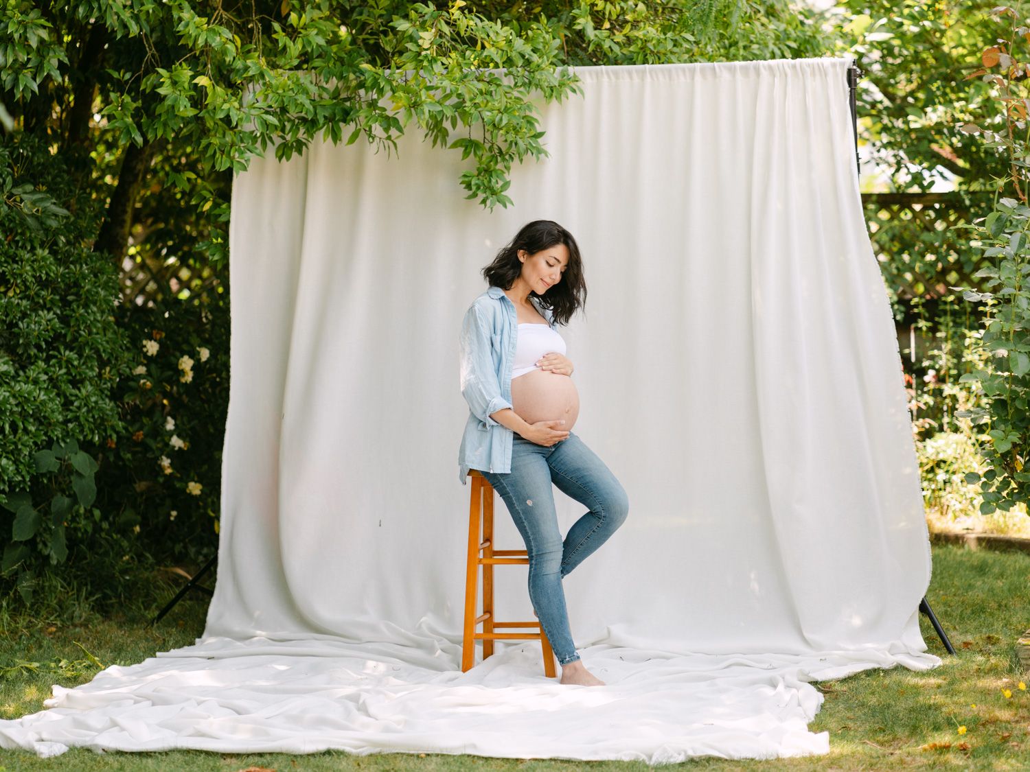Pregnant woman in blue shirt and jeans posing outdoors with baby bump, sitting on stool with garden backdrop.
