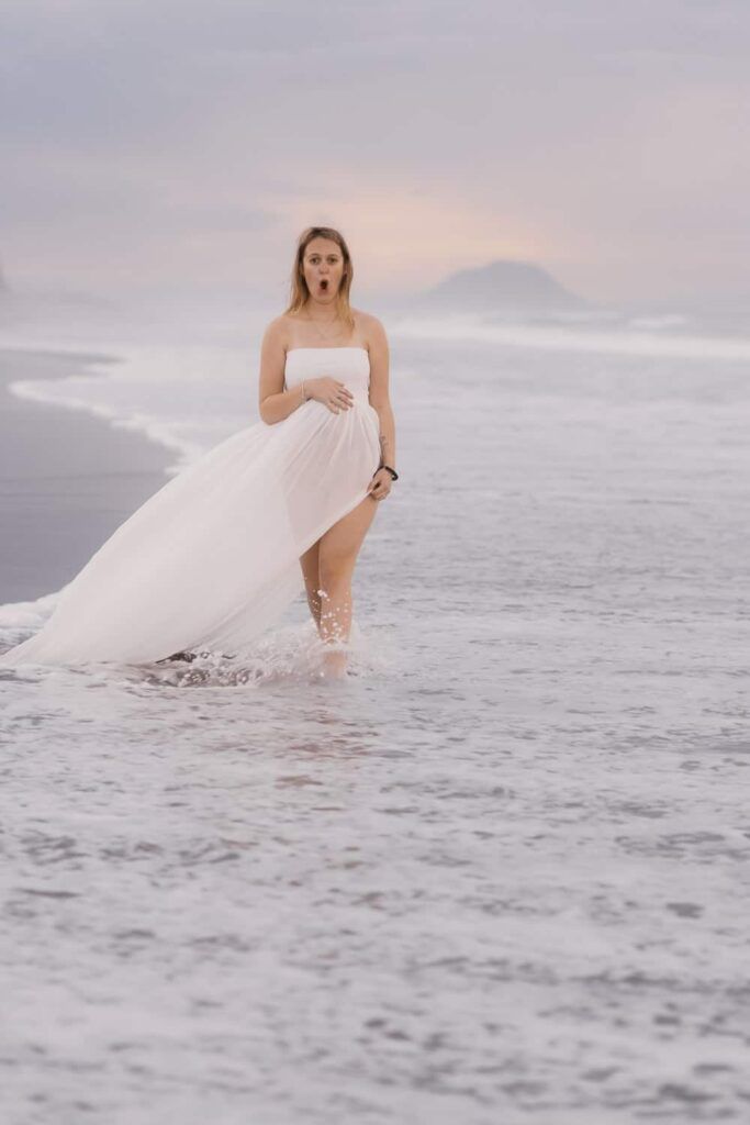 Pregnant woman in white dress walking in ocean waves at sunset beach.