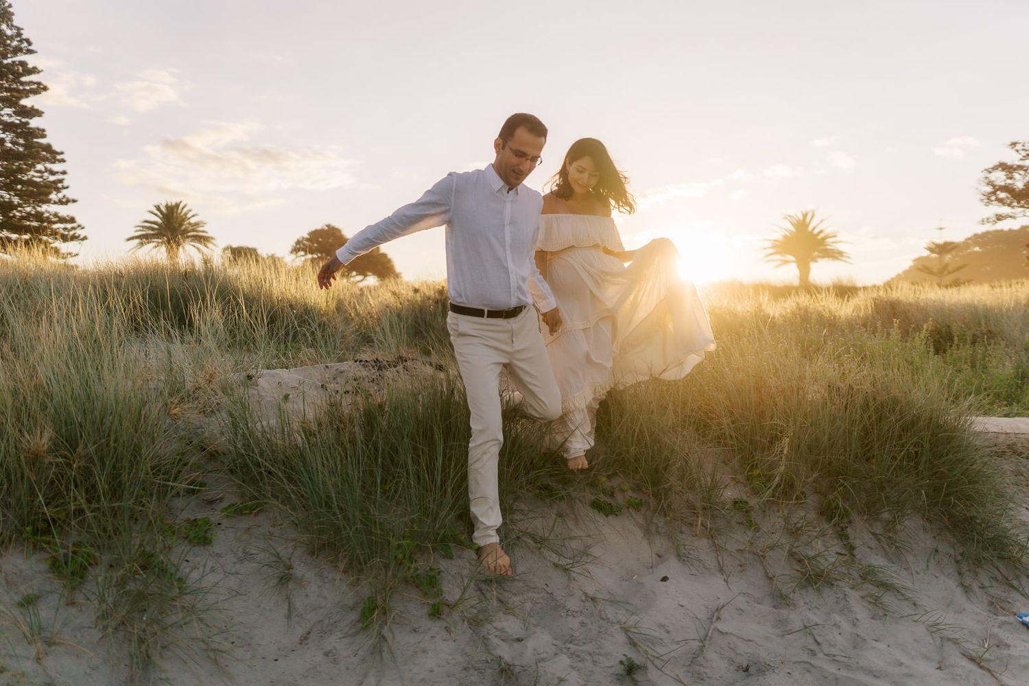 Couple holding hands running on sandy beach dunes at golden sunset with palm trees in background.