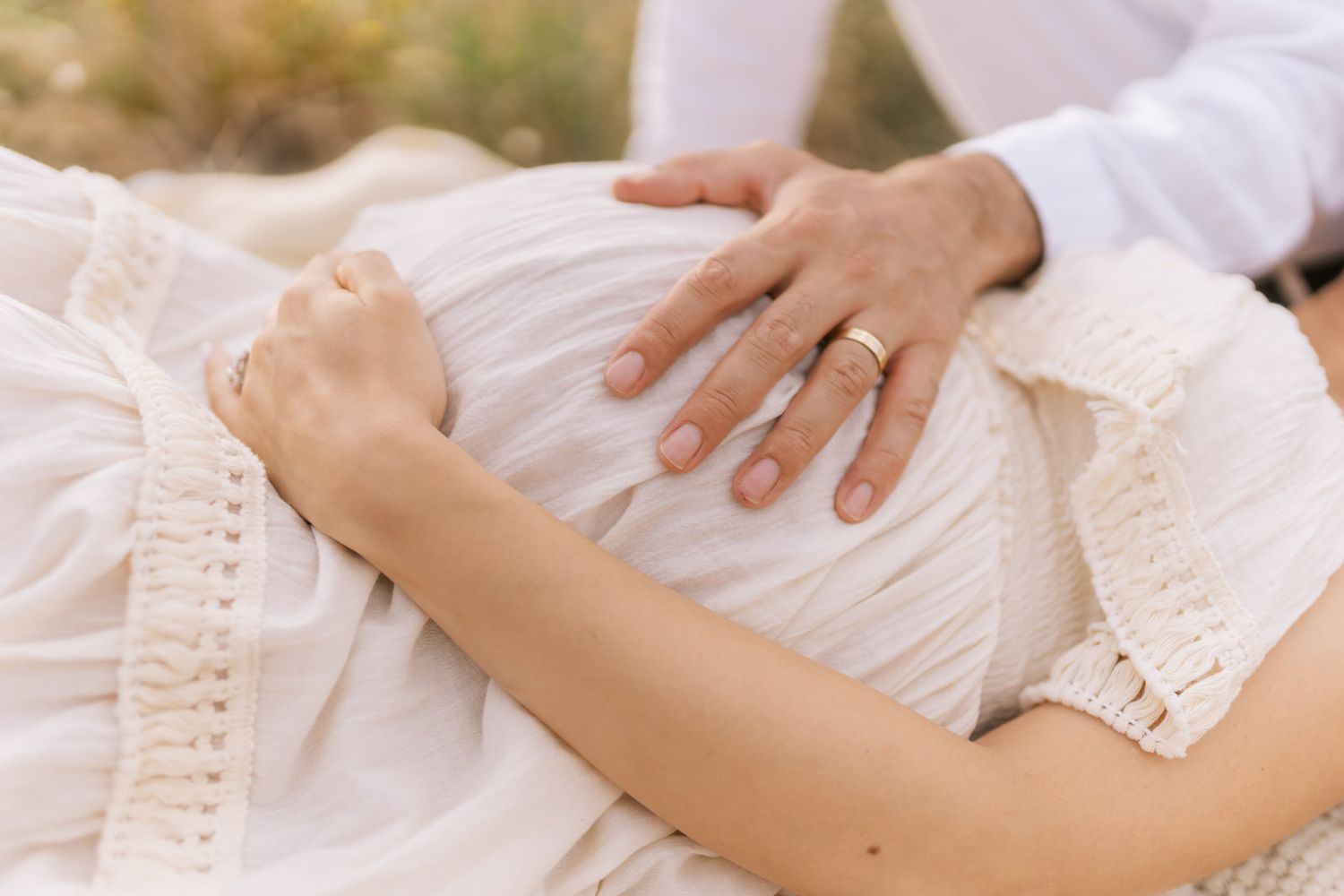 Couple tenderly touching pregnant belly outdoors, wearing wedding rings, in soft white dress during maternity photo session.