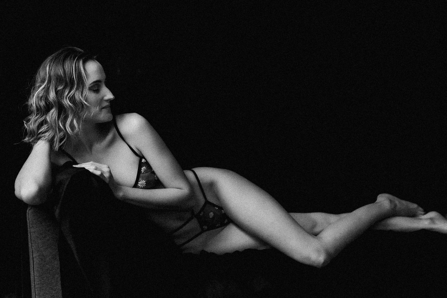Black and white boudoir photo of a blonde woman reclining gracefully in lingerie against a dark background.