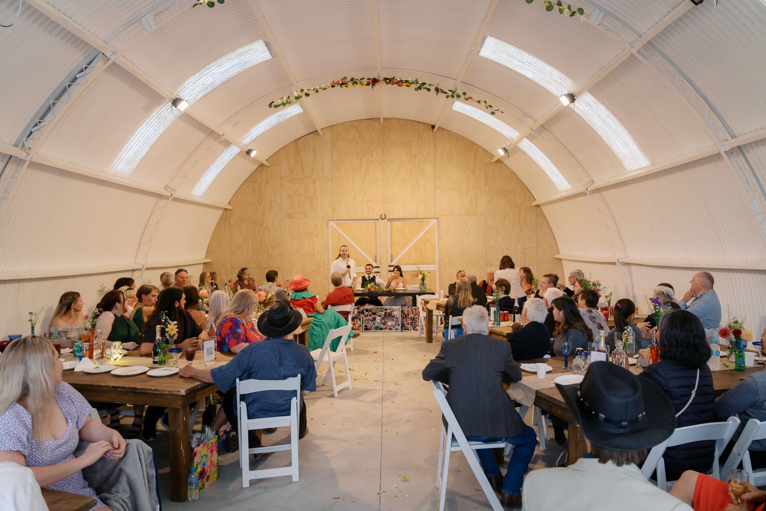 Guests gather at long wooden tables inside a curved white tent venue decorated with floral garlands for a group dining event.