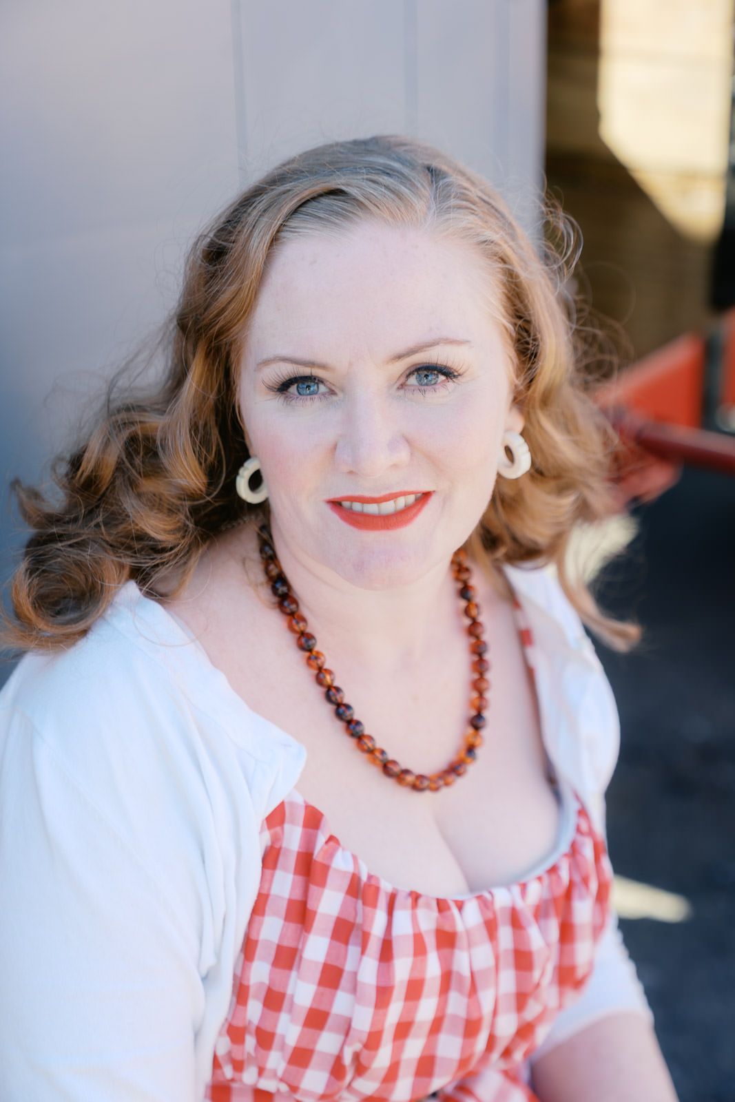 Smiling woman with red lipstick, gingham dress, amber necklace, and hoop earrings poses outdoors in natural light.