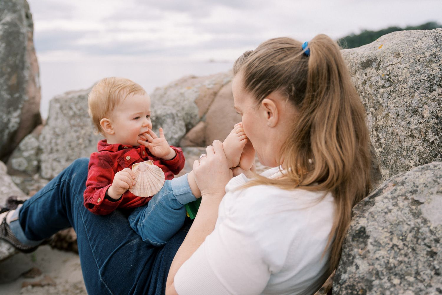 A young mother sits against rocky boulders outdoors, lovingly interacting with her smiling baby dressed in red.