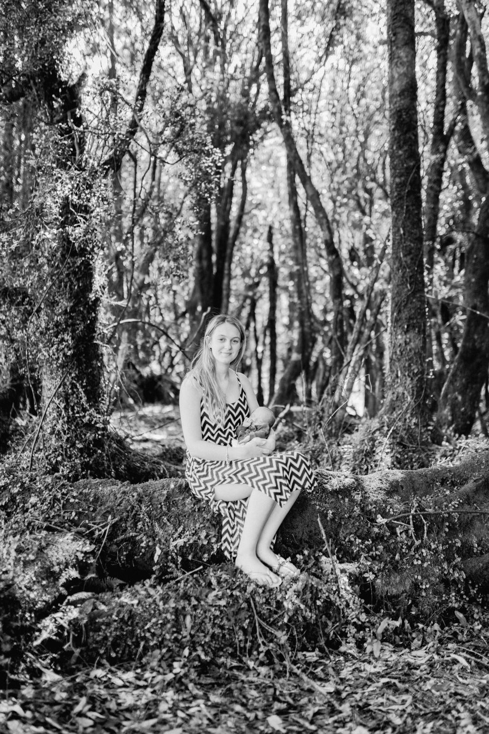 Black and white photo of a young woman in a chevron dress sitting on a tree root in a lush forest setting.
