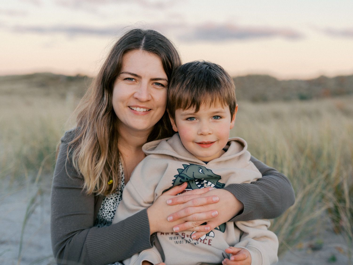Mother and young son posing together on sandy beach dunes at sunset, boy holding toy dinosaur.