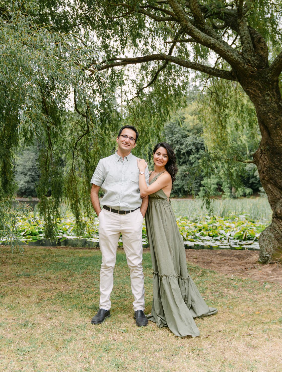 Couple posing under a willow tree in a lush garden, man in khaki pants and woman in sage green maxi dress.