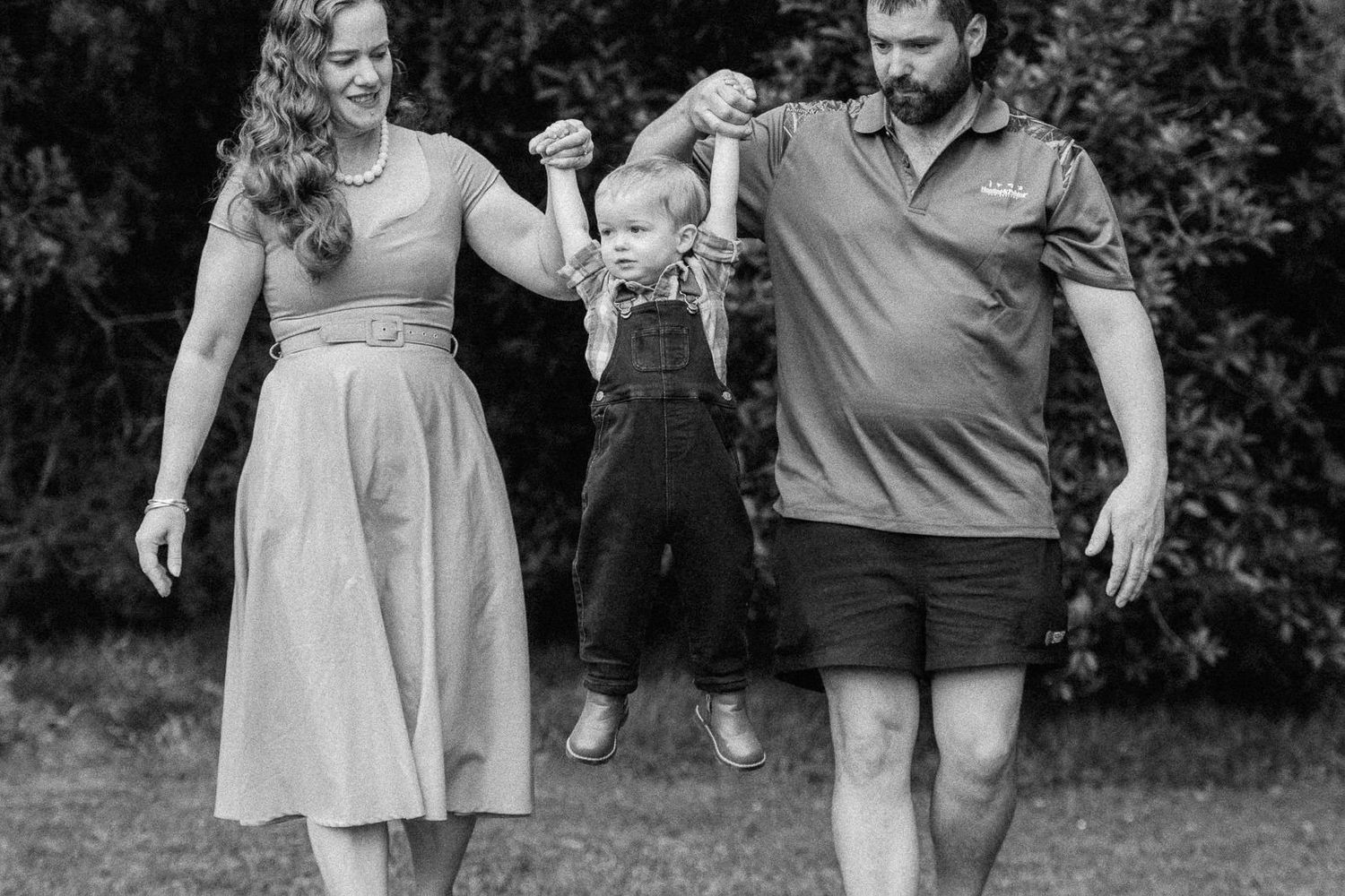 Black and white photo of a happy family with parents swinging their toddler between them outdoors.