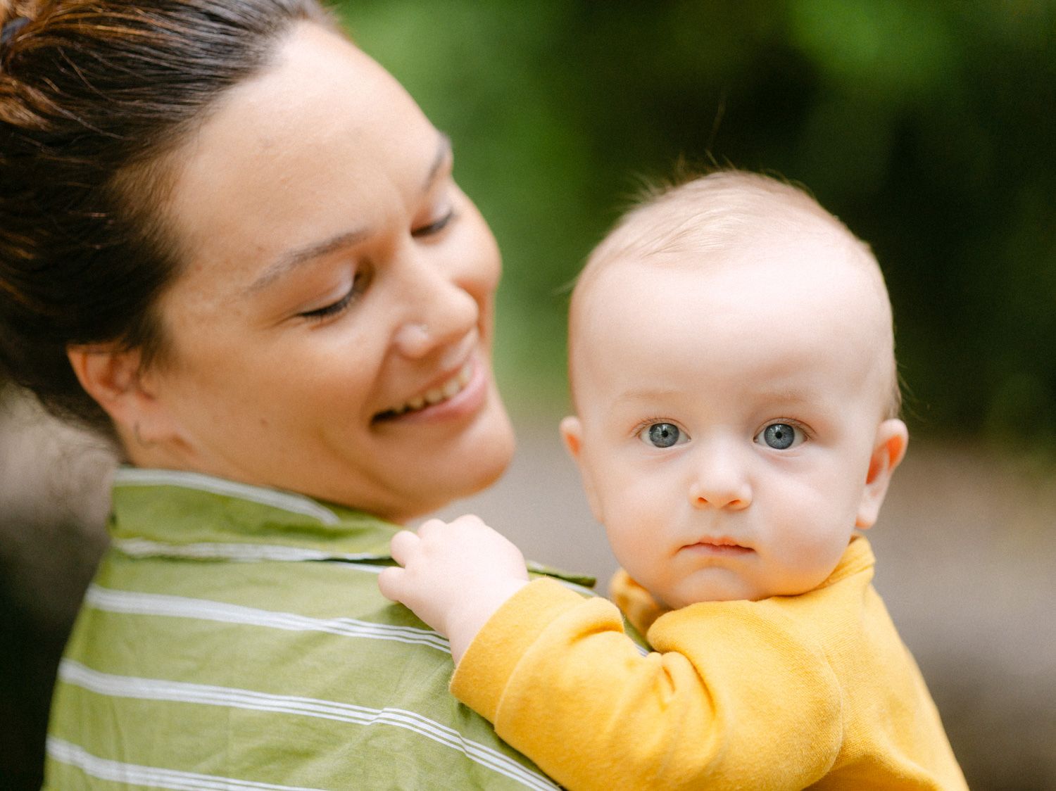 Smiling mother holding her blue-eyed baby outdoors, both dressed in green and yellow, sharing a tender moment.