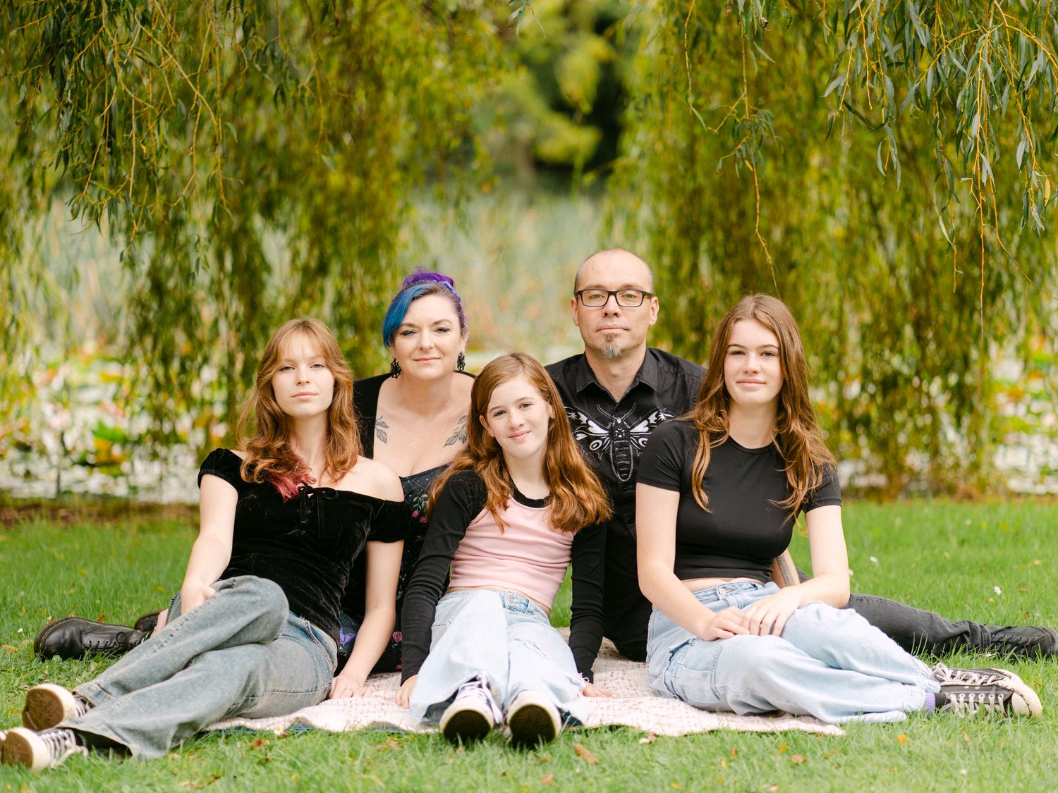 Family of five sitting on grass under a willow tree in a lush green park for an outdoor portrait.