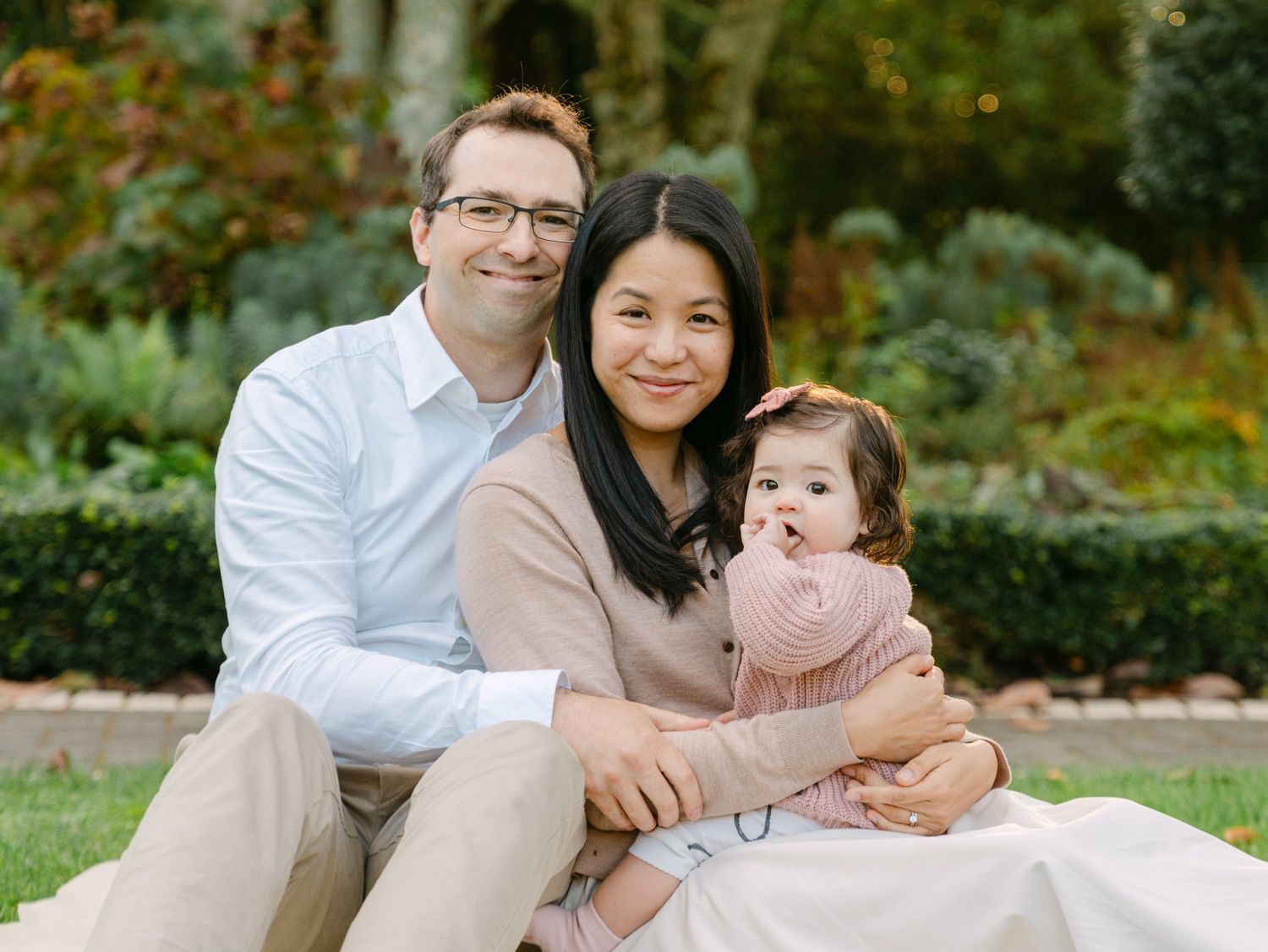 Happy family portrait with couple and baby girl sitting on grass in a lush garden setting.