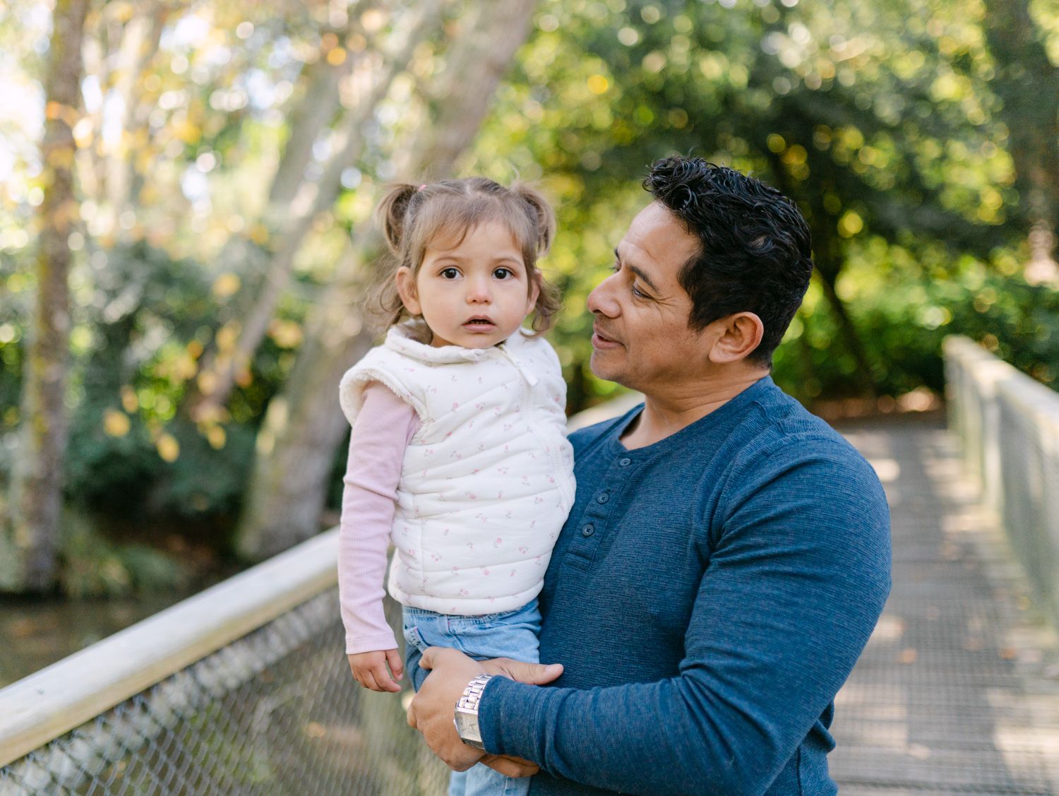 A man holding a toddler girl on a wooden bridge surrounded by lush green trees in a park.
