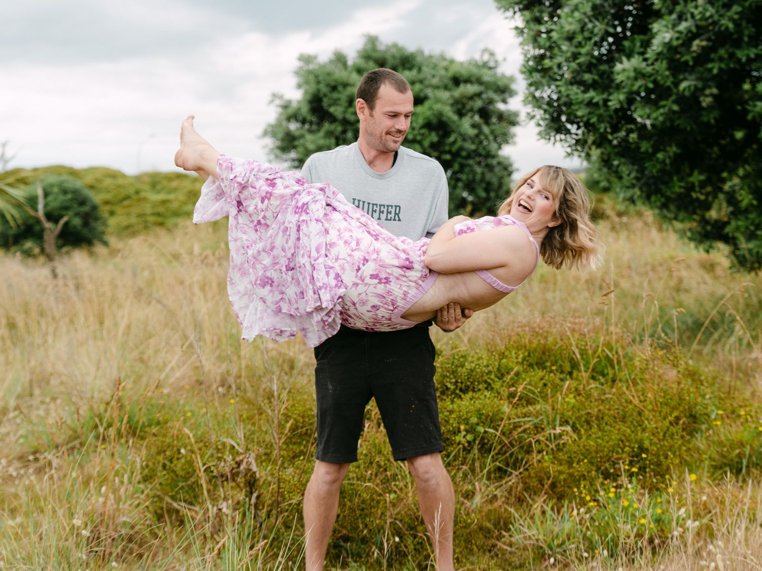 A smiling man lifts a laughing woman in a floral dress in a lush green meadow.
