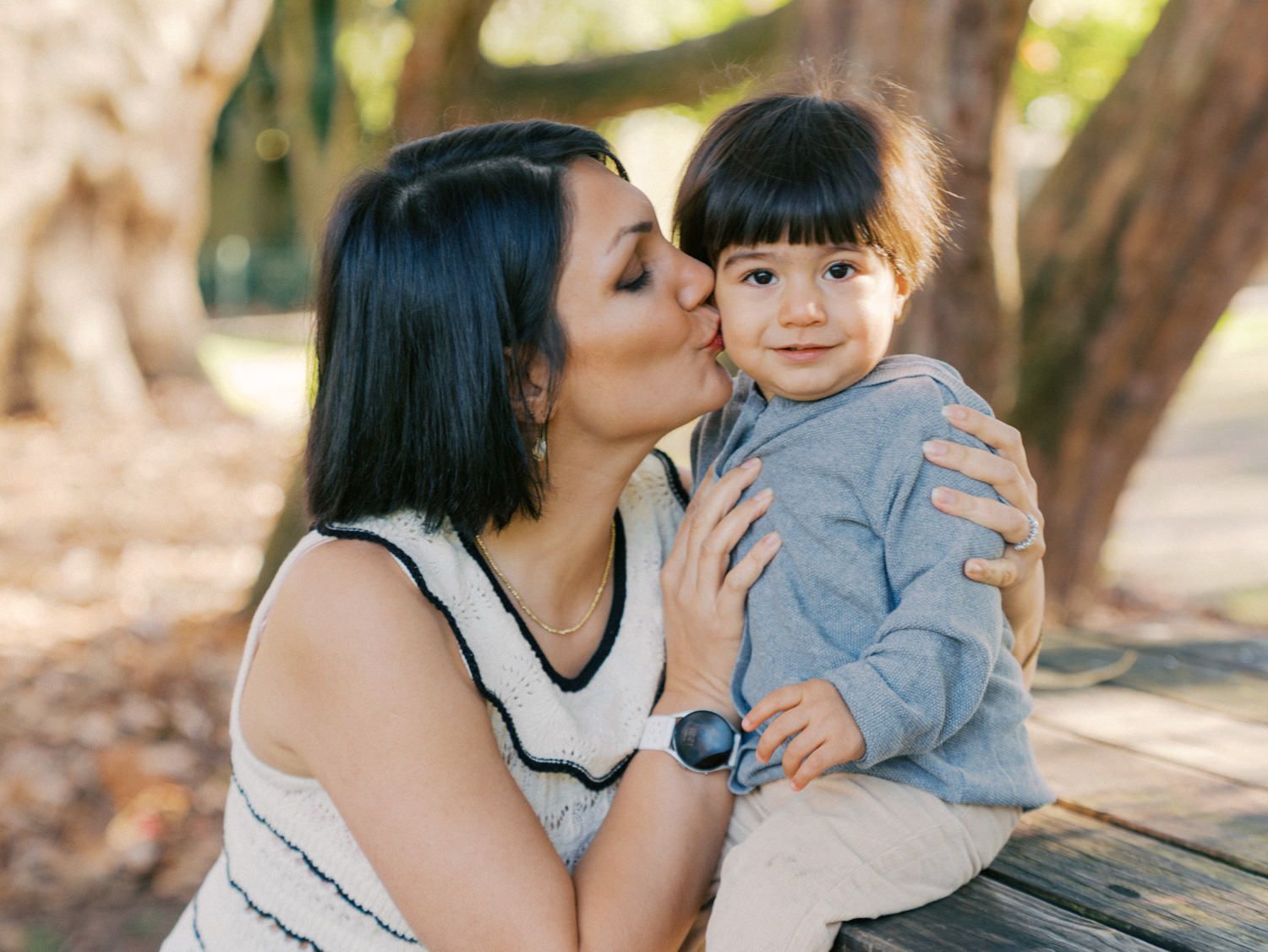 A mother tenderly kisses her toddler on the cheek outdoors in a sunlit park setting.