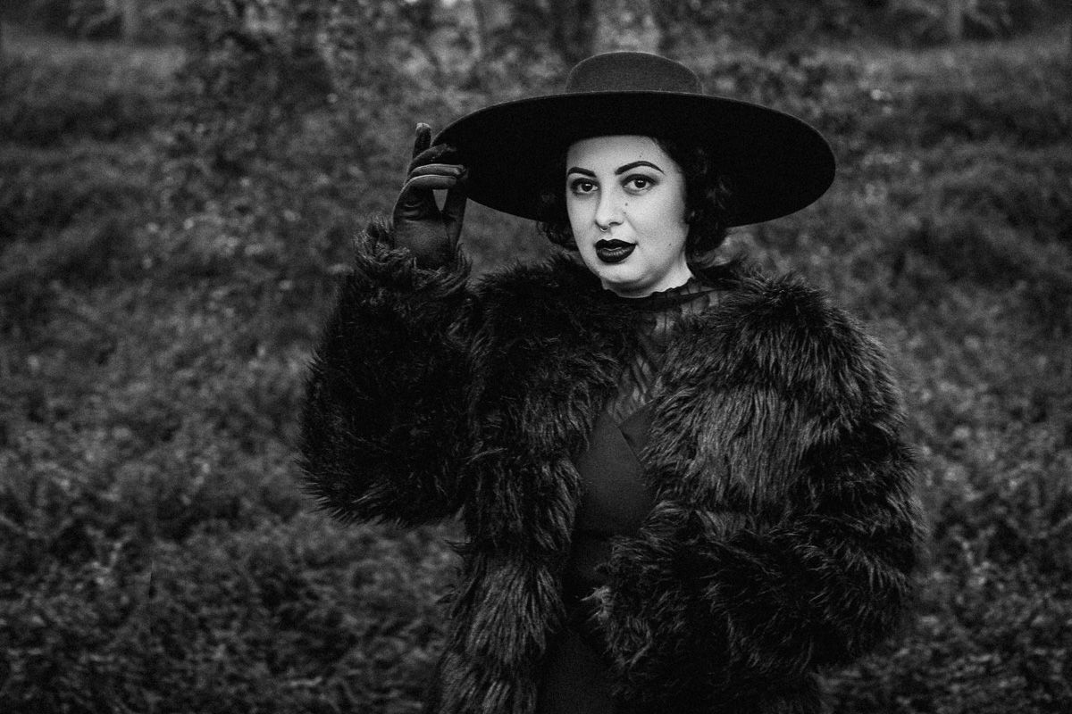 Black and white portrait of a woman in fur coat and wide-brim hat posing outdoors.