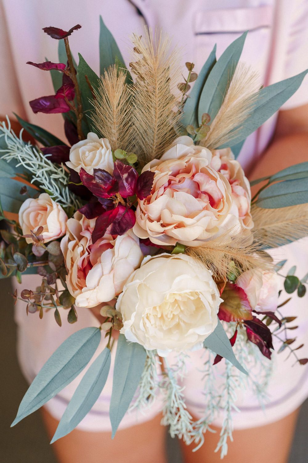 Boho bridal bouquet with blush peonies, white roses, burgundy blooms, pampas grass, and eucalyptus held by woman in pink dress.