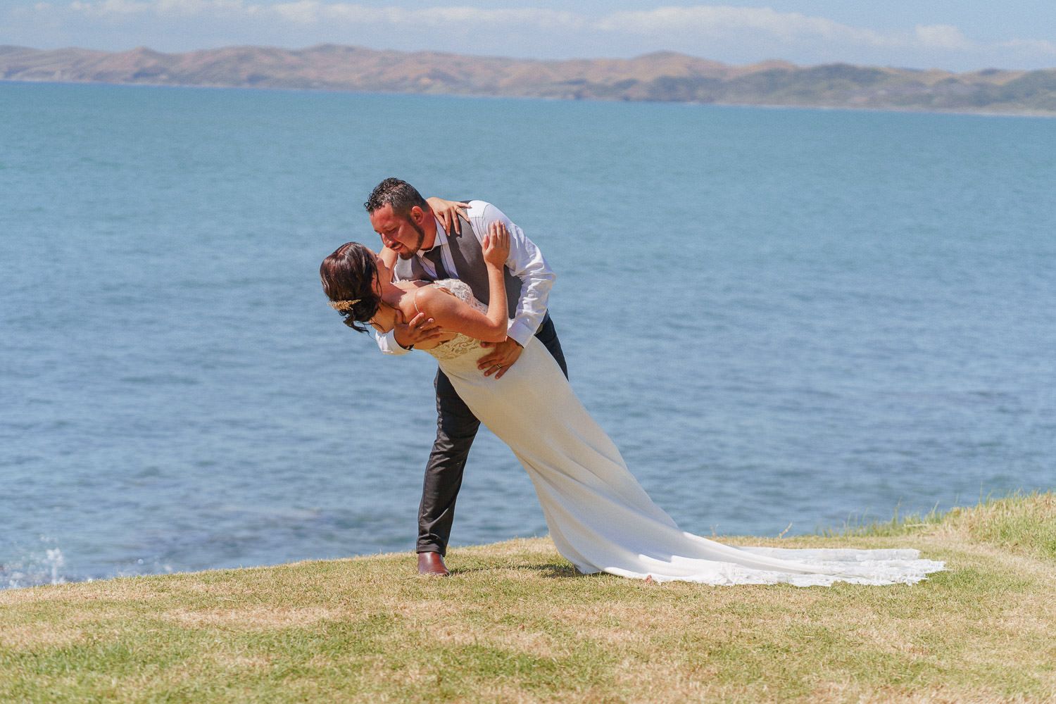 Bride and groom sharing a romantic dip kiss on a grassy cliff overlooking a scenic blue bay on their wedding day.