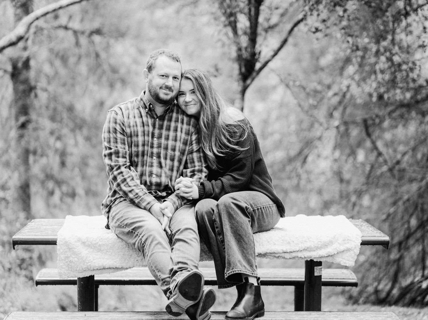 Couple smiling together on a wooden bench outdoors in a black and white engagement photo among autumn trees.
