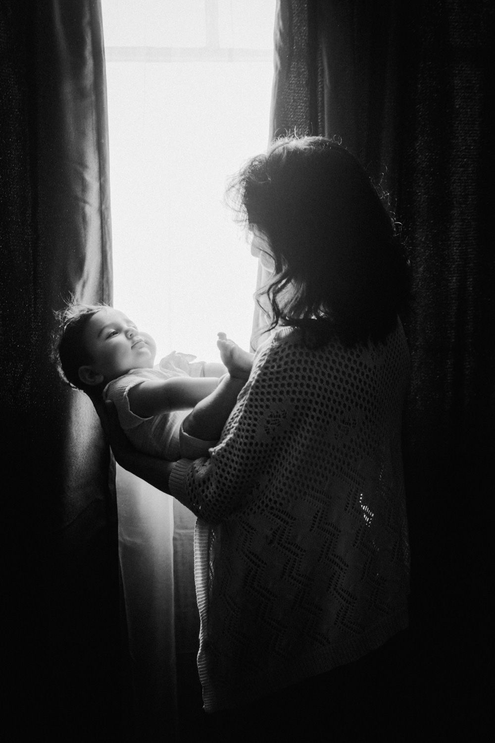 Black and white photo of a mother holding her newborn baby by a bright window with curtains.