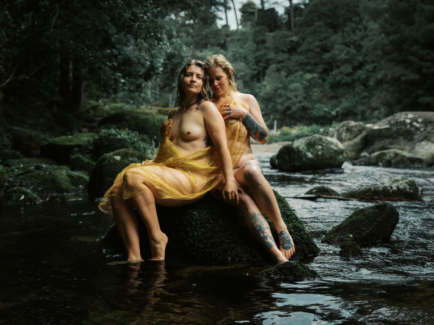 Two women in golden yellow dresses sit on rocks by a forest stream surrounded by lush green trees.