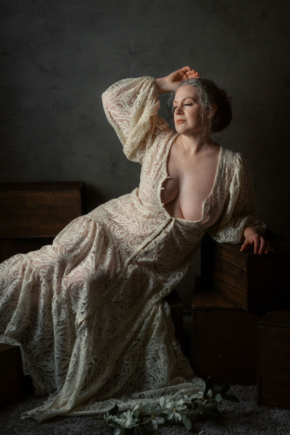 A woman in a flowing lace gown poses gracefully on a wooden chest in a moody, dimly lit studio setting.