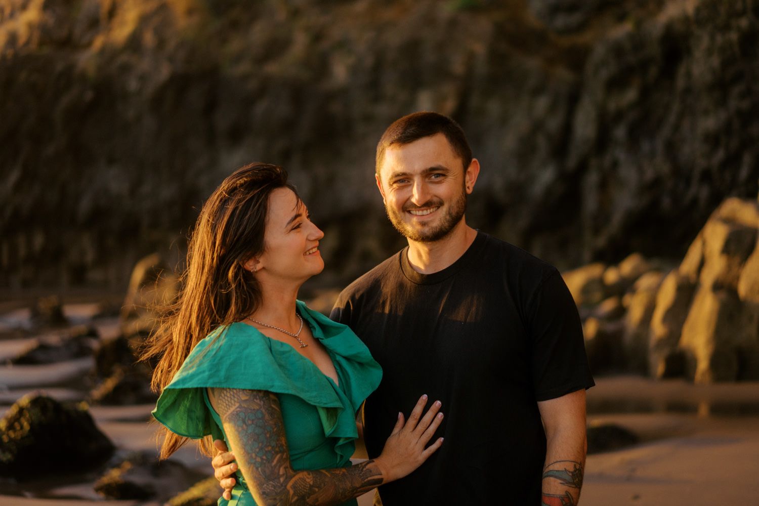 Couple smiling together on a rocky beach at golden hour, woman in green top with tattoos, man in black shirt.