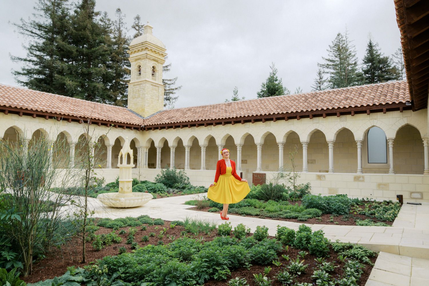 Woman in red dress twirling in a Mediterranean-style courtyard with arched colonnade and lush gardens.