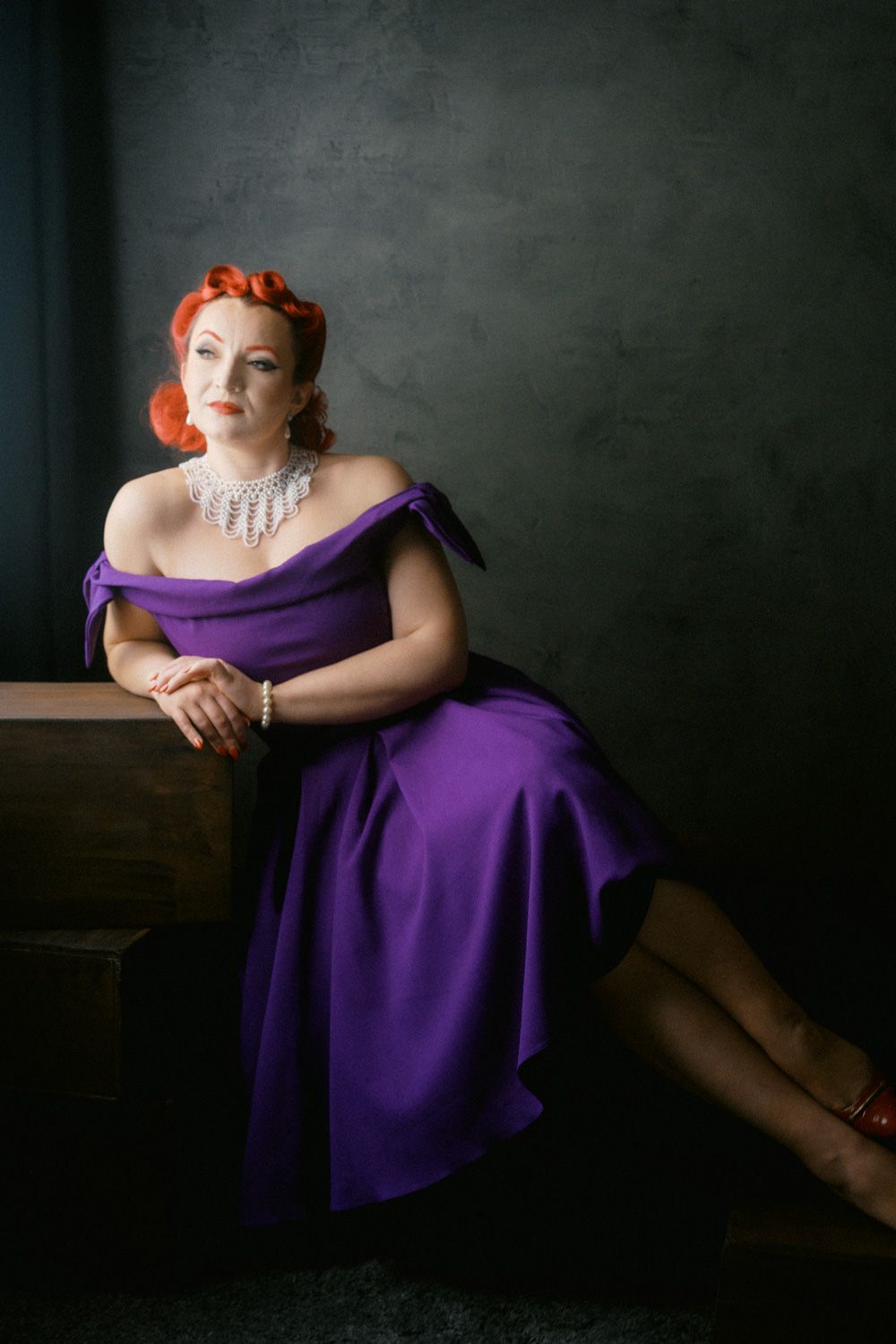 Woman in purple off-shoulder dress with red hair and pearl necklace sits elegantly against a dark gray wall.