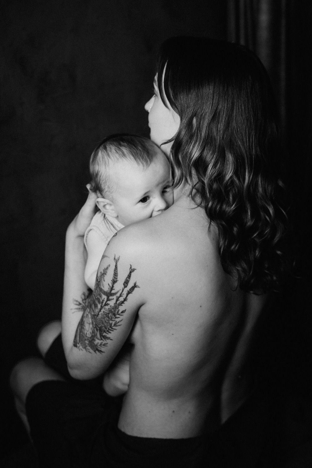 Black and white photo of a mother with a floral back tattoo breastfeeding her newborn baby in soft studio lighting.