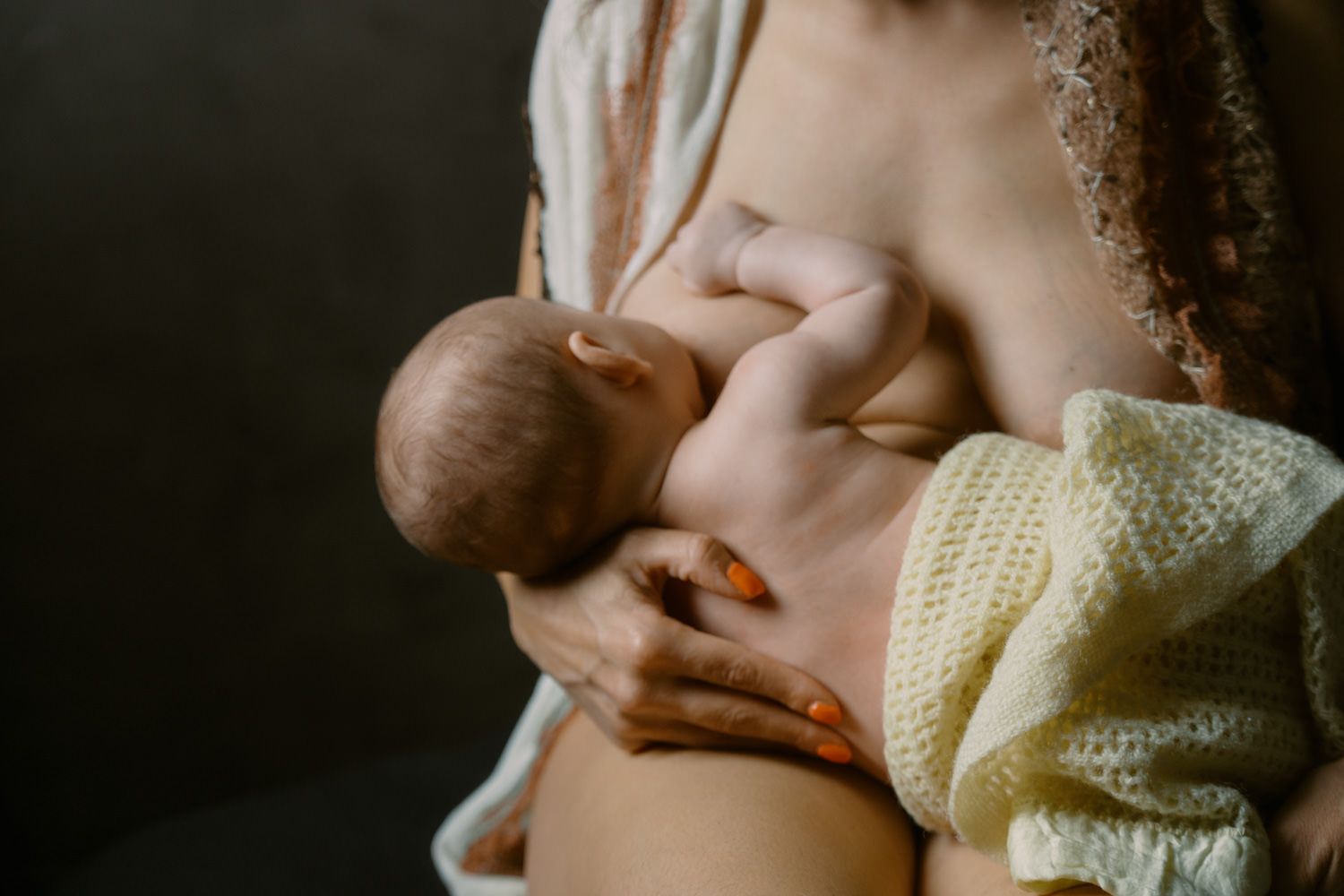 Mother breastfeeding newborn baby, cradling infant in arms while wearing a yellow knit sweater.