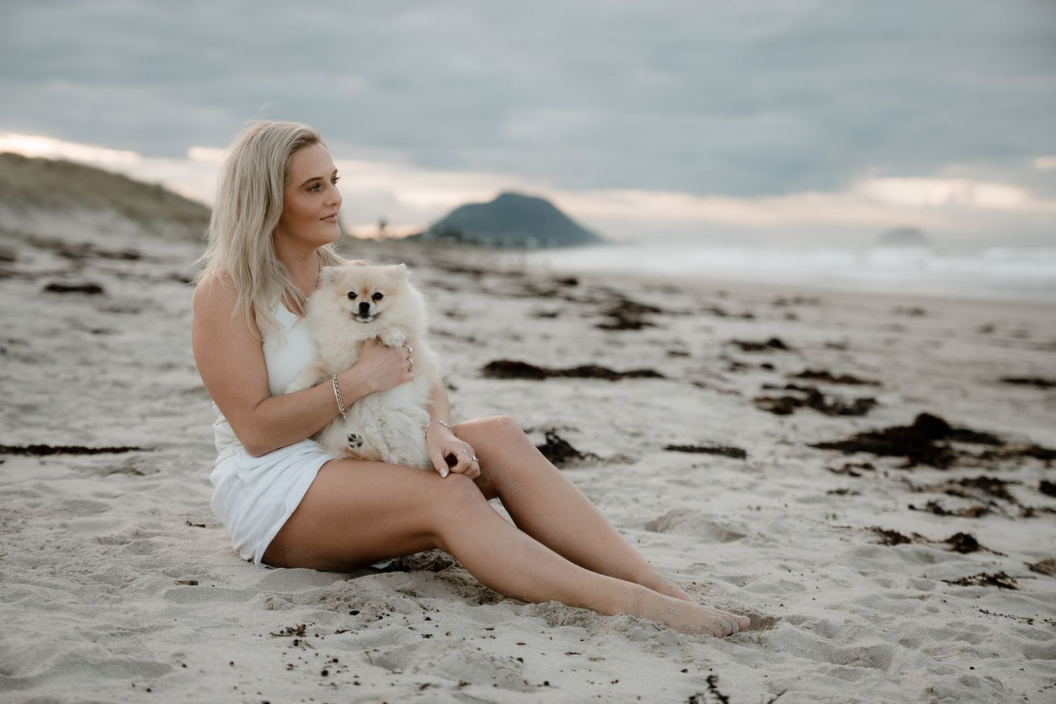Blonde woman in white dress sitting on sandy beach holding a small white Pomeranian dog with cloudy sky backdrop.