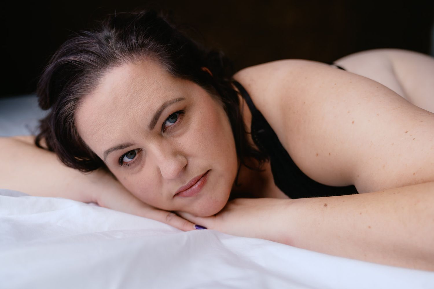 Woman in black lingerie lying on white bed, resting her chin on her hands, gazing at camera.