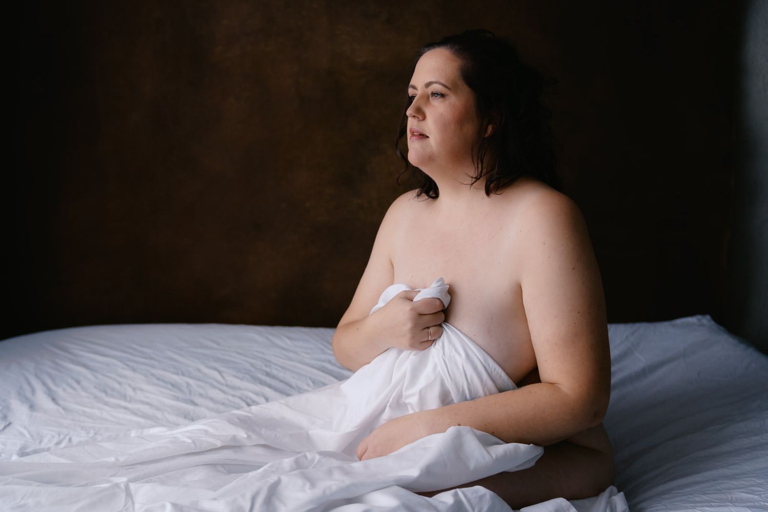 A woman sits on a white bed holding a sheet, gazing thoughtfully in a moody, dimly lit boudoir setting.