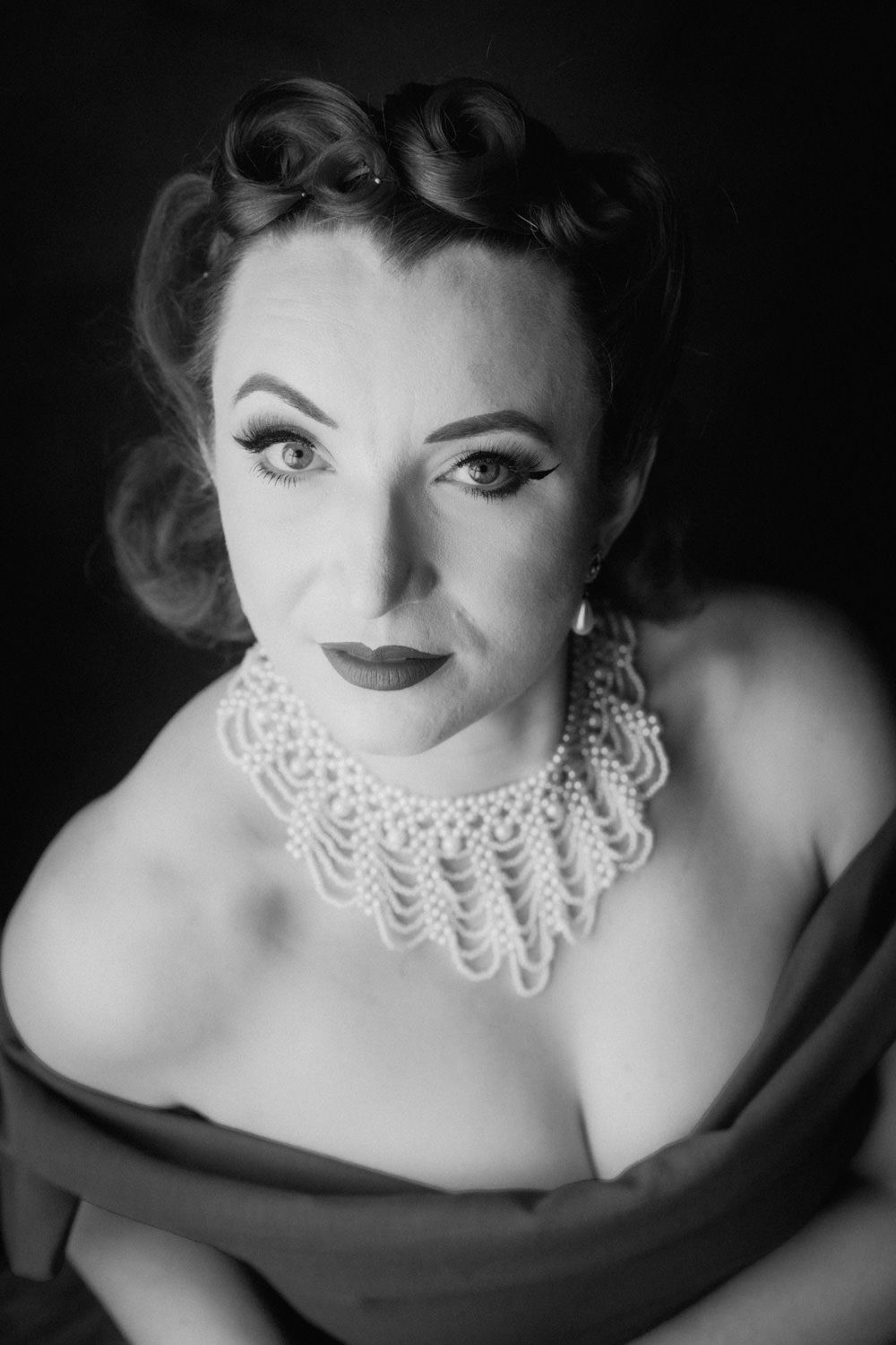 Black and white vintage portrait of a woman with retro hairstyle wearing a pearl necklace and off-shoulder dress.