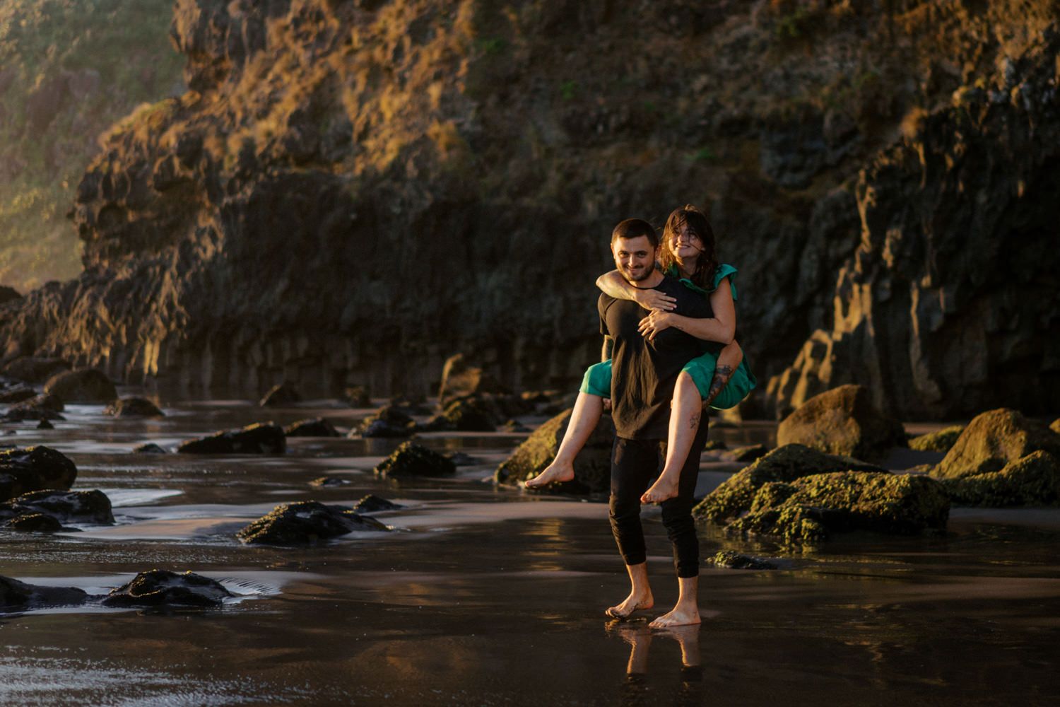 Couple sharing a romantic piggyback moment on a rocky beach shore at golden hour.