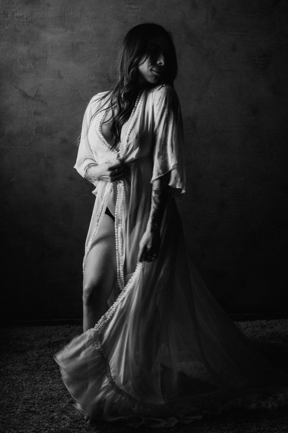 Black and white studio portrait of a woman in a flowing white robe gown posing dramatically against a dark textured wall.