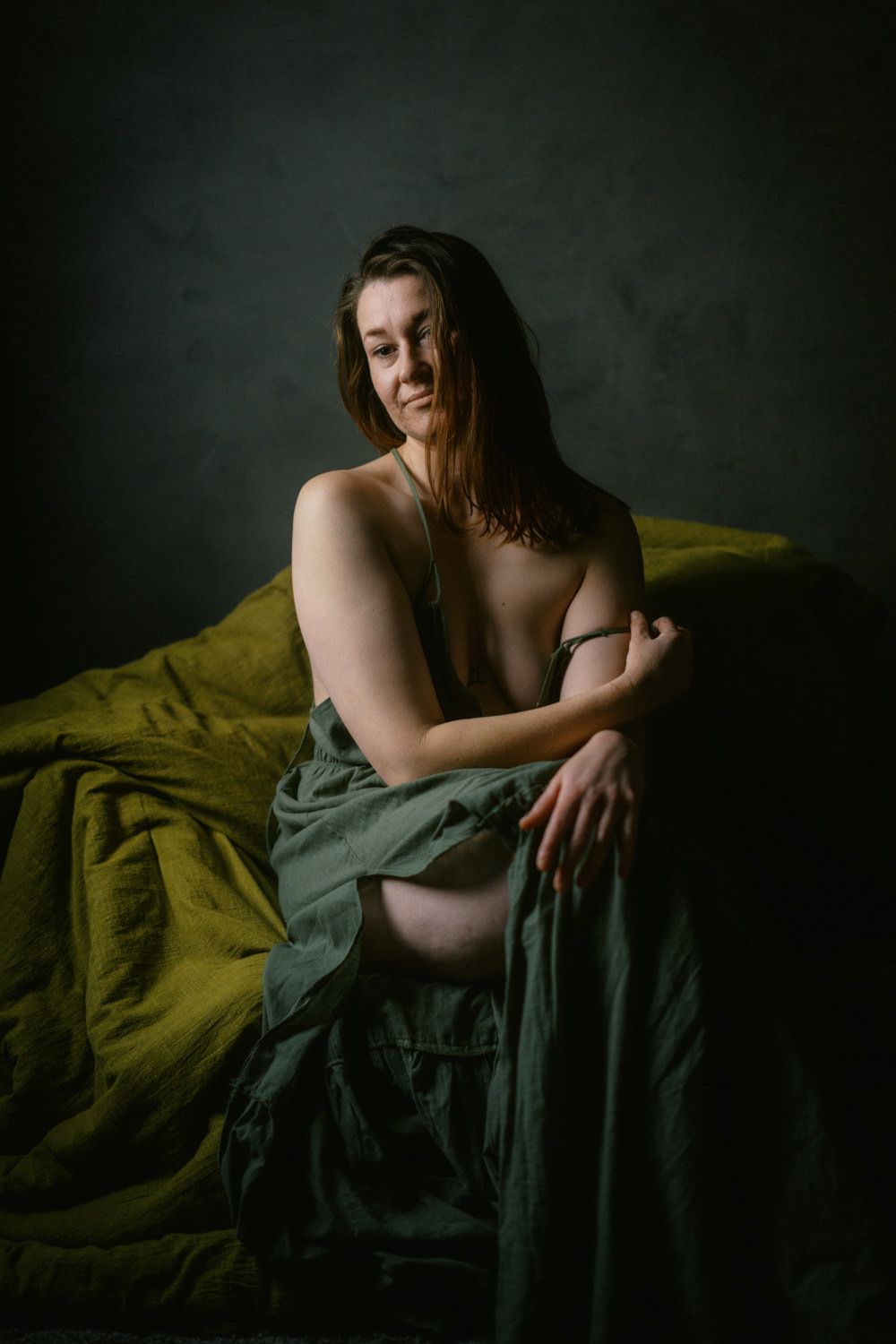 A woman sits wrapped in olive green fabric against a dark backdrop in a moody fine art portrait.