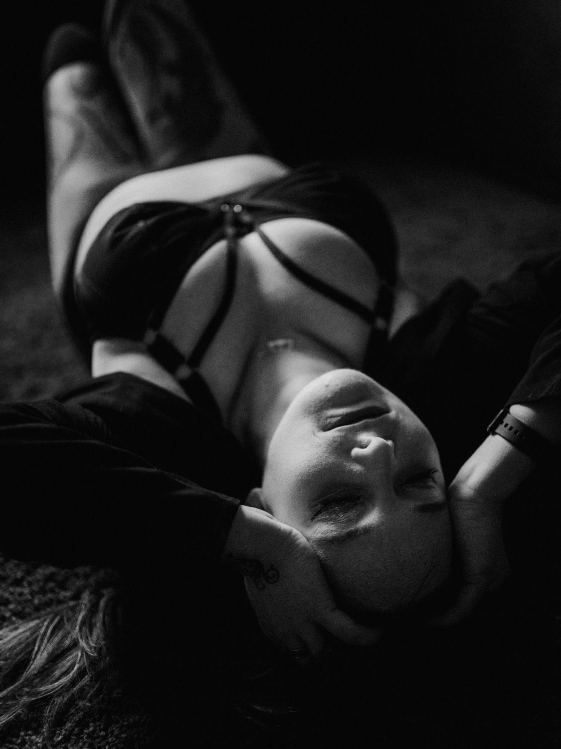 Black and white boudoir photo of a woman lying down in lingerie with dramatic shadows and moody lighting.