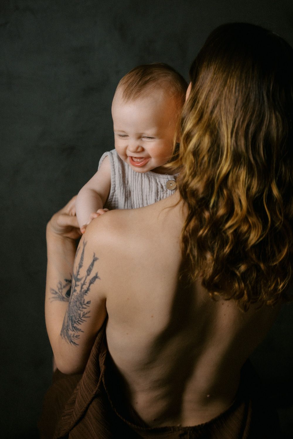 A tattooed mother holds her laughing baby against a dark studio backdrop.